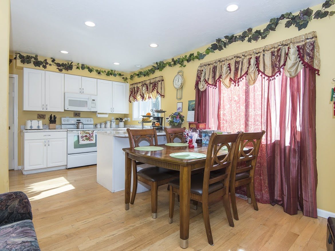 A cozy kitchen and dining area with wooden floors, featuring a dining table, white cabinets, and decorative curtains.
