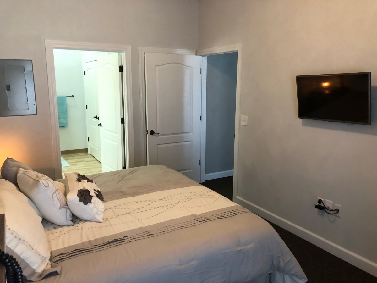 A minimalist bedroom with a bed, wall-mounted television, and open doors leading to an adjoining room.