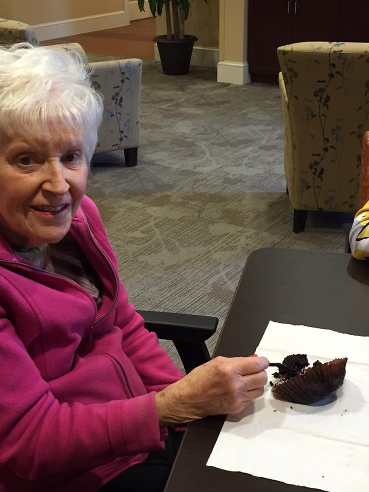 An elderly woman in a pink jacket is enjoying a piece of chocolate cake while sitting at a table indoors.