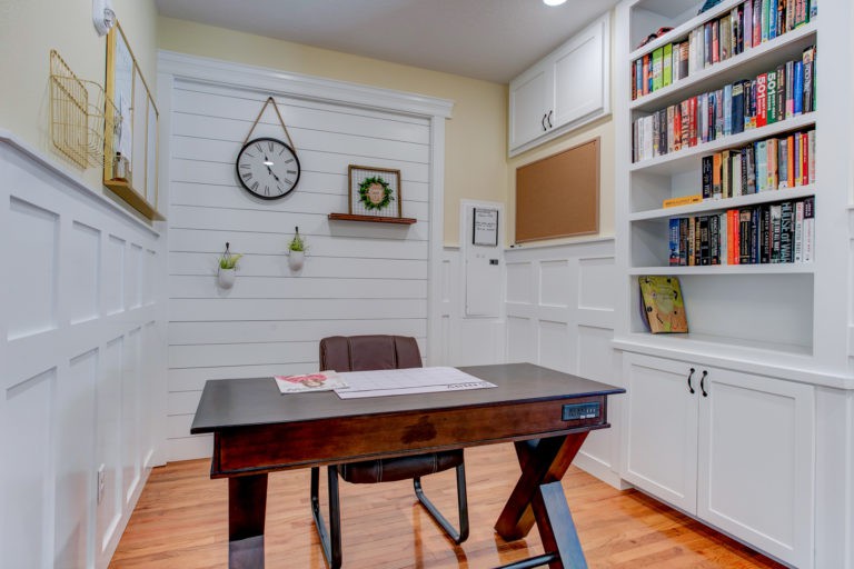 A cozy home office with a wooden desk, chair, and bookshelves, decorated with a clock and wall decor.
