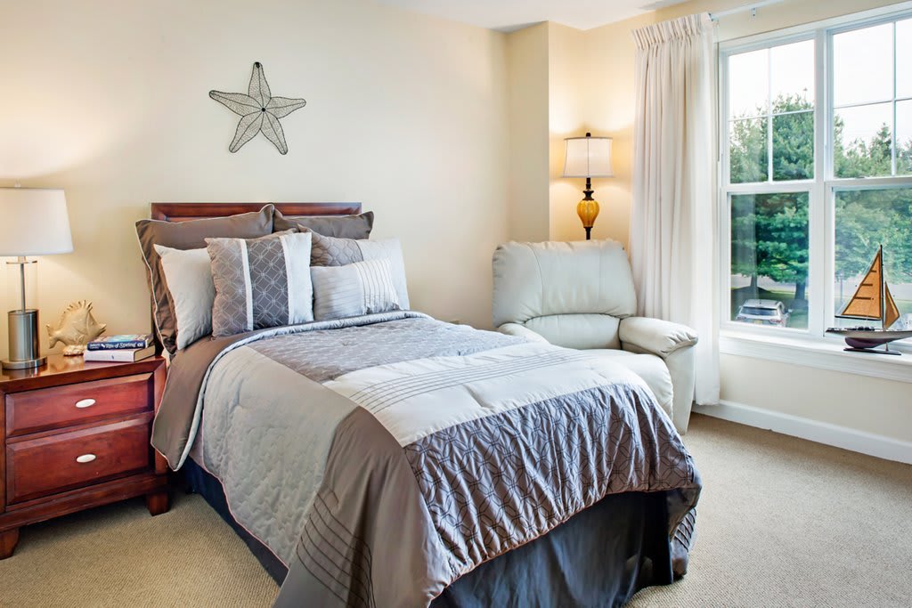 A cozy bedroom with a neatly made bed, a comfortable chair, and natural light coming through the window.