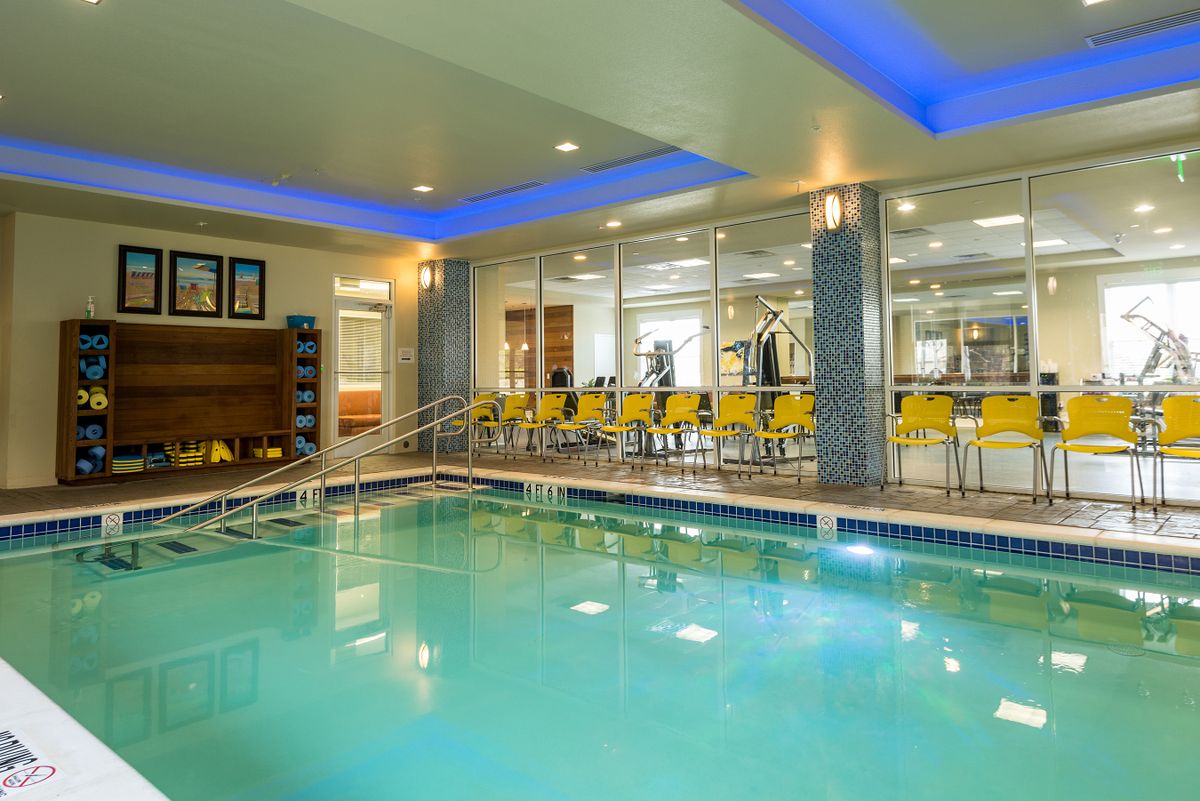 A modern indoor swimming pool with a seating area and glass walls leading to a fitness center.