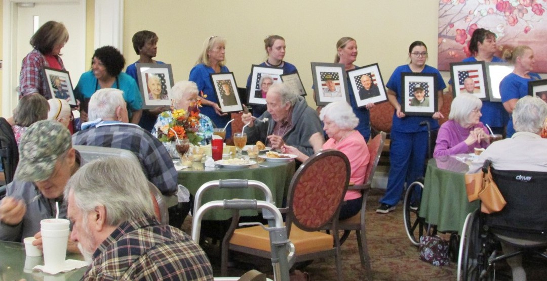 A group of people at a community event hold portraits while elderly participants enjoy a meal.