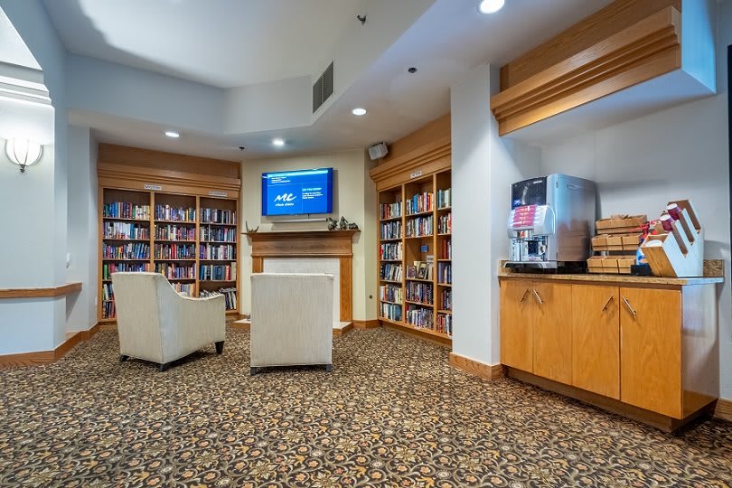 A cozy library setting with bookshelves, comfortable seating, a television, and a coffee machine.