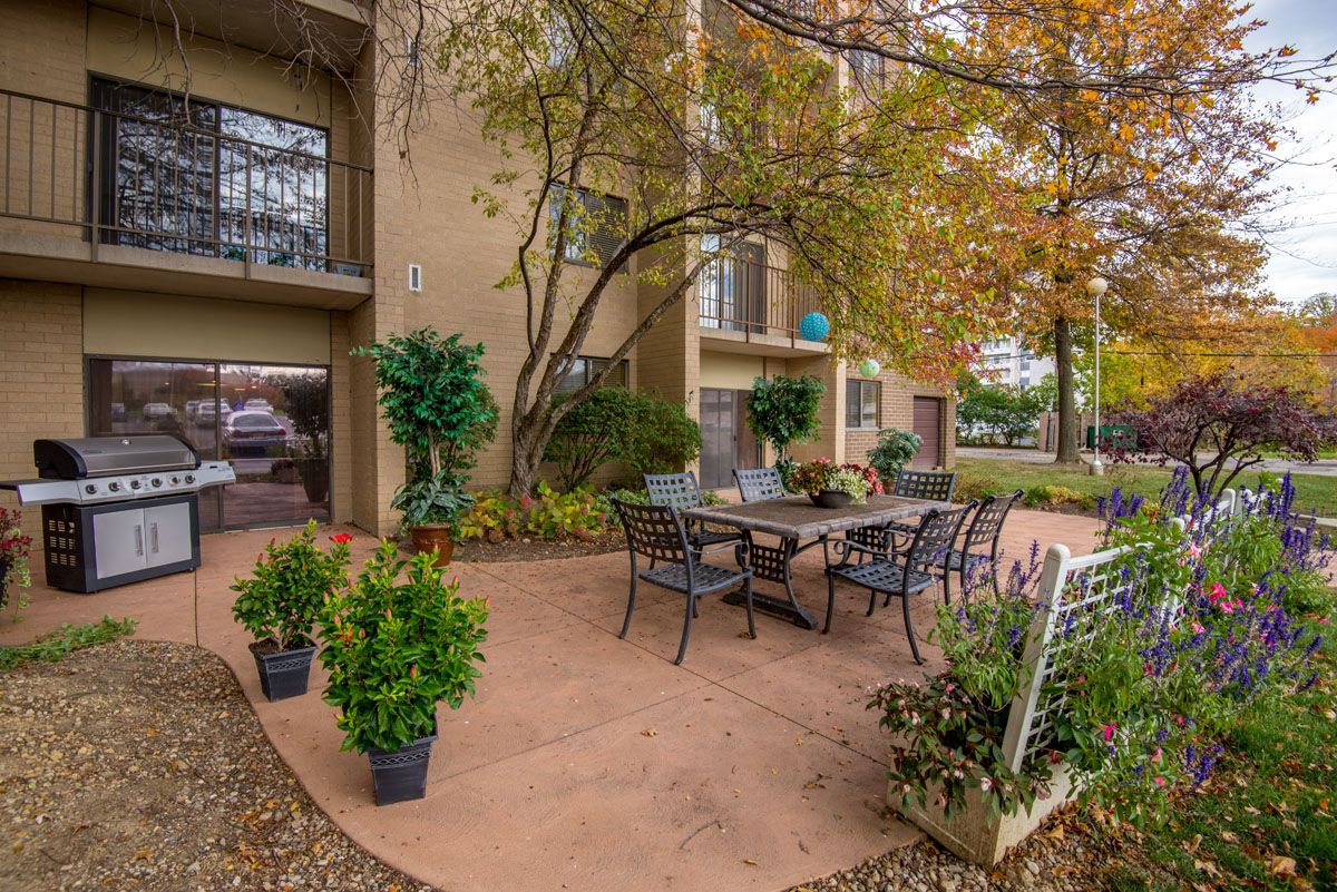 A cozy outdoor patio area with seating, surrounded by greenery and an apartment building in the background.
