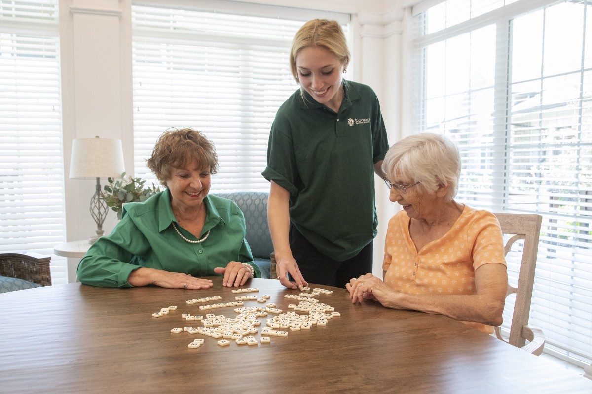 A caregiver assists two senior women with a game at a table indoors.