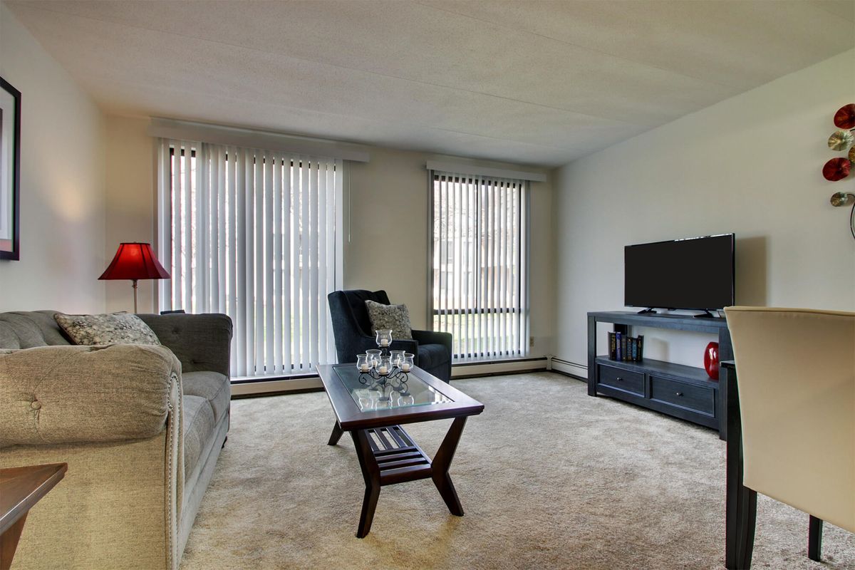 A cozy living room featuring a sofa, armchair, coffee table, and TV with large windows and vertical blinds.