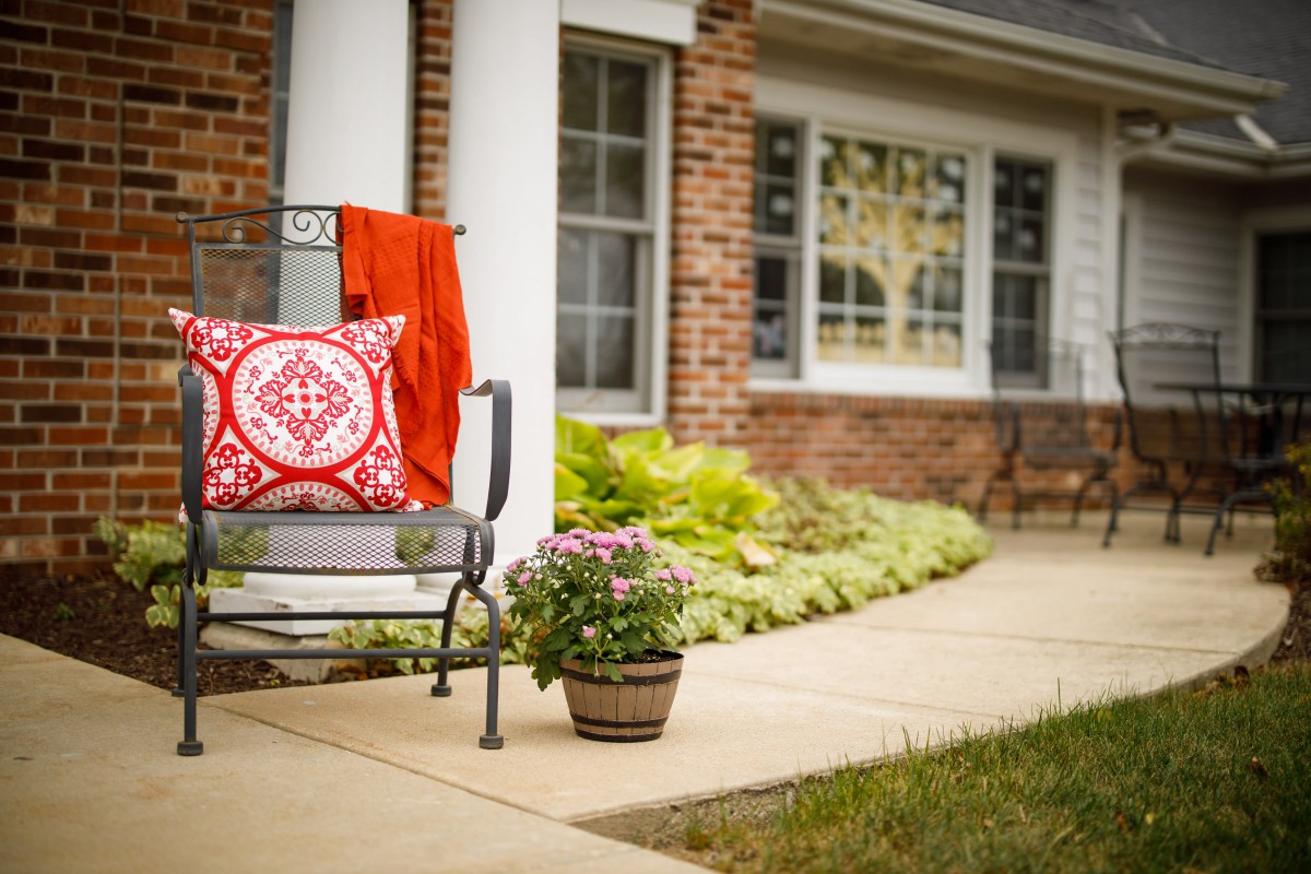 A cozy patio bench with a patterned pillow and potted plant against a brick house facade.