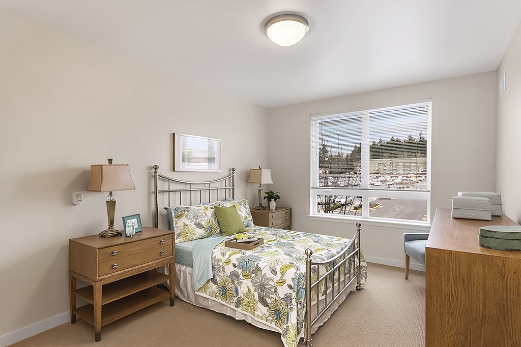 A cozy bedroom with a metal frame bed, floral bedding, and wooden furniture, illuminated by natural light from a large window.