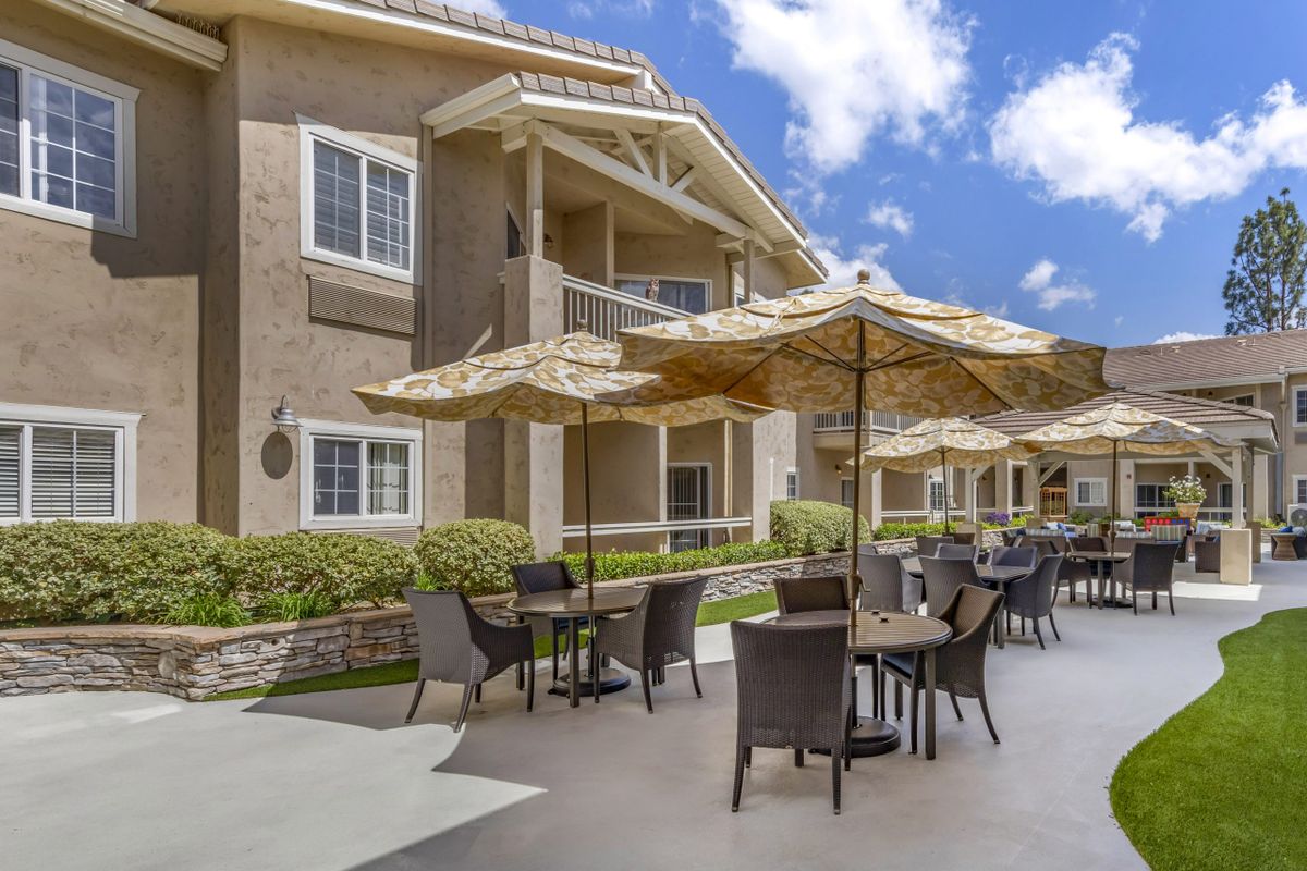 Outdoor seating area with tables and umbrellas in front of an apartment building on a sunny day.