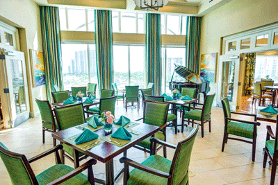 A brightly lit dining area with green chairs, neatly set tables, and a grand piano by large windows.