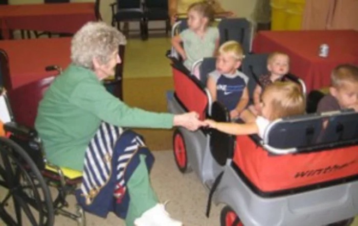 An elderly woman interacts with children seated in a stroller inside a daycare setting.