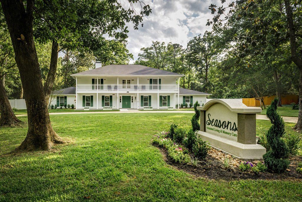 A colonial-style house surrounded by a lush green lawn and trees, with a sign in the foreground under a cloudy sky.