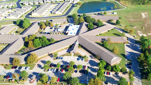 An aerial view of a building complex surrounded by trees and a pond, featuring a spacious parking lot.