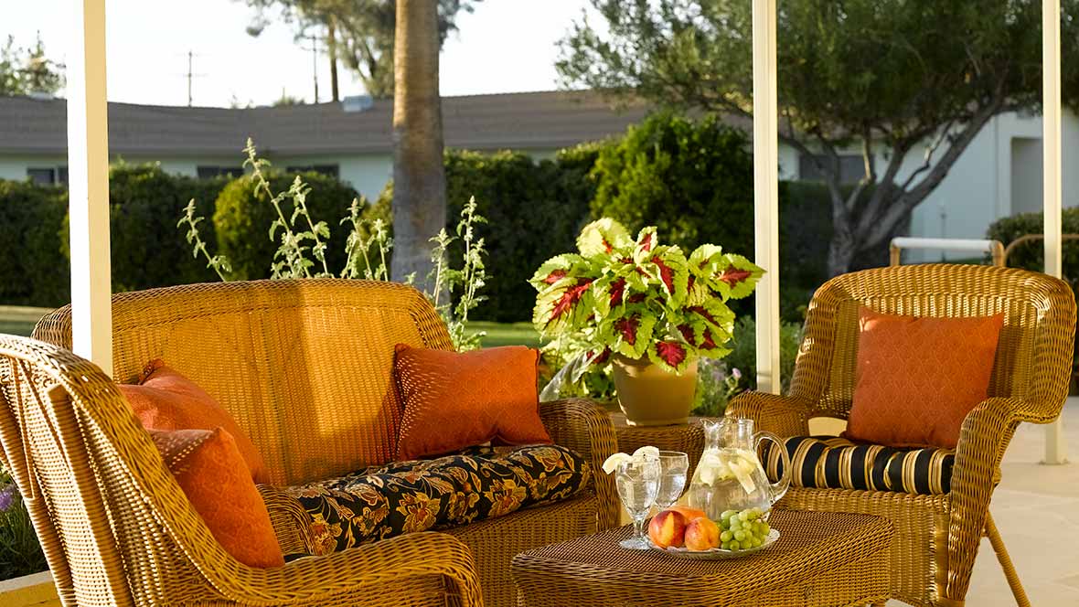 A cozy outdoor patio setting with wicker chairs, colorful cushions, and a table set with refreshments and plants.