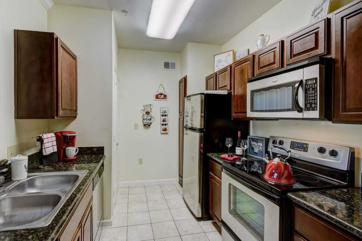 A compact kitchen with wooden cabinets, appliances including a microwave, oven, and fridge, along with tiled flooring.