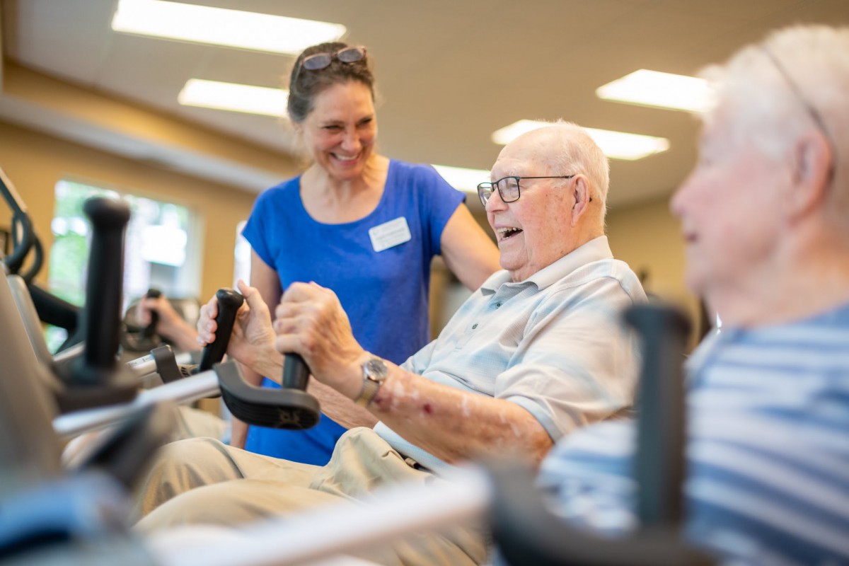 An elderly man exercising in a gym with assistance from a trainer.