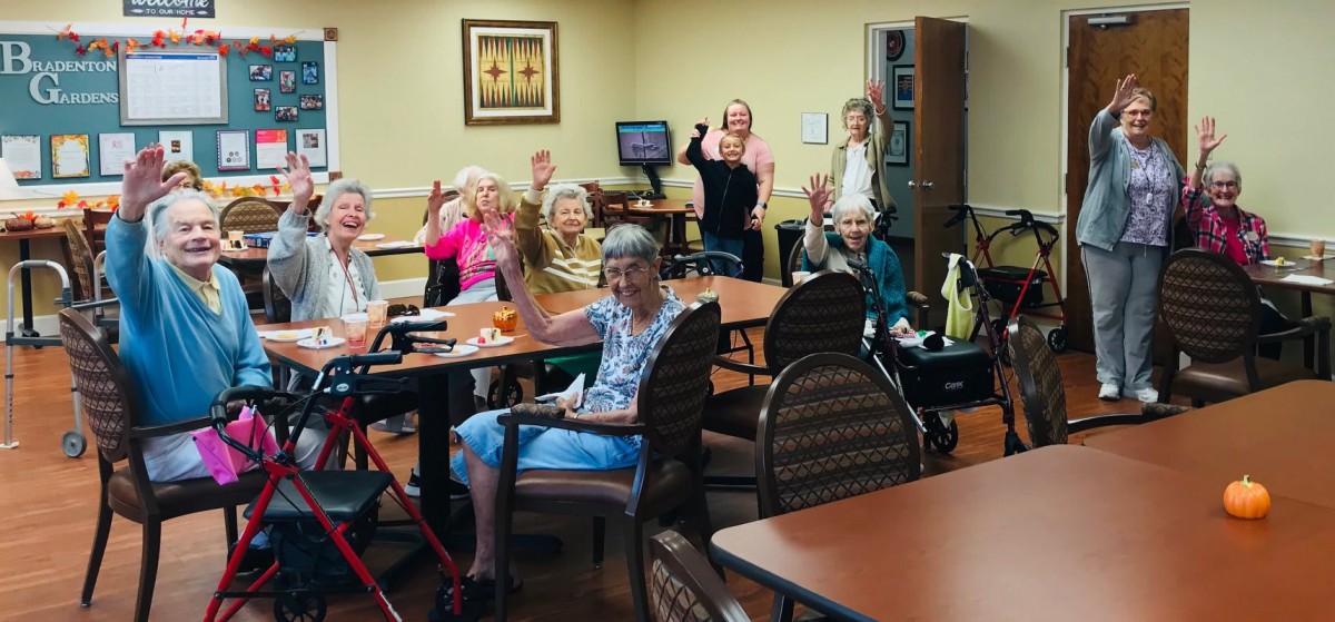 A cheerful group of seniors gathered indoors, waving and smiling.