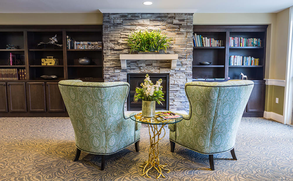 Cozy living room with armchairs facing a stone fireplace, surrounded by bookshelves and decorated with greenery.