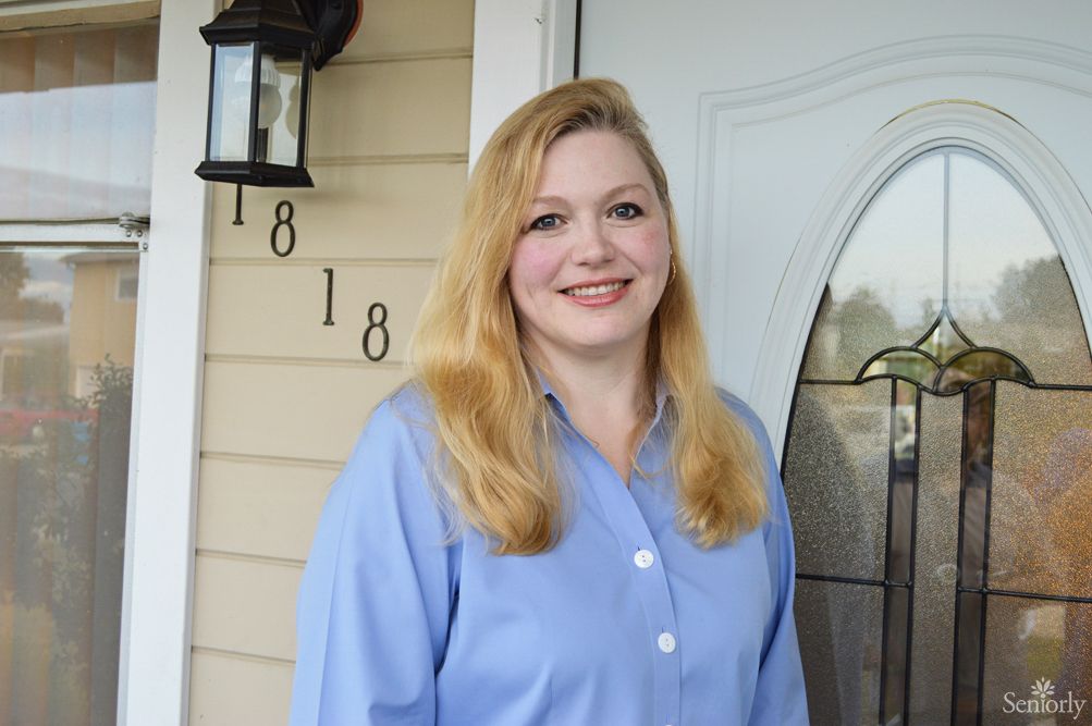 A smiling woman with blonde hair in a blue shirt stands outside a house.