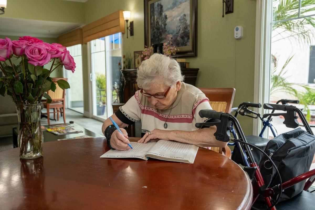 An elderly person sitting at a table, writing in a book with a walker nearby.
