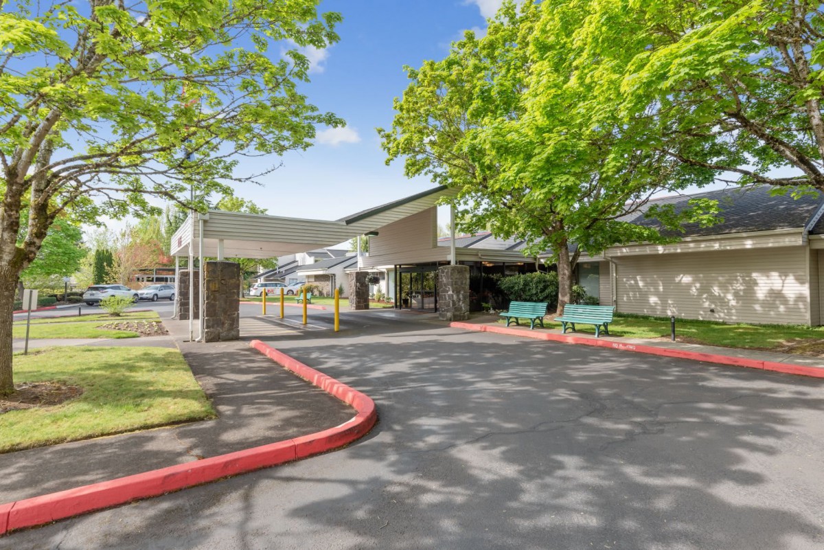 A sunny day view of the entrance to a building surrounded by trees and a parking lot.