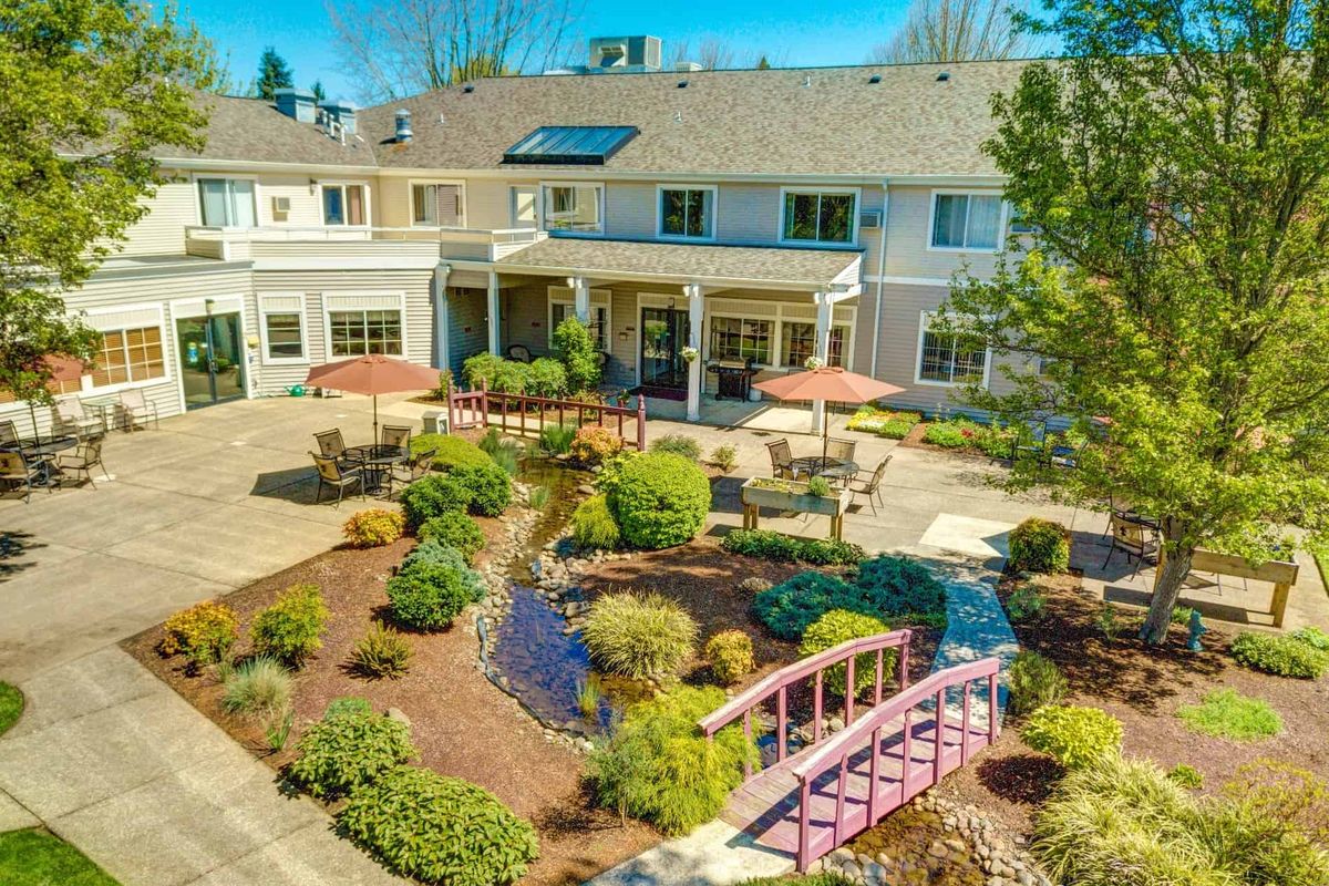 A beautifully landscaped garden with a small bridge and patio area in front of a large residential building on a sunny day.