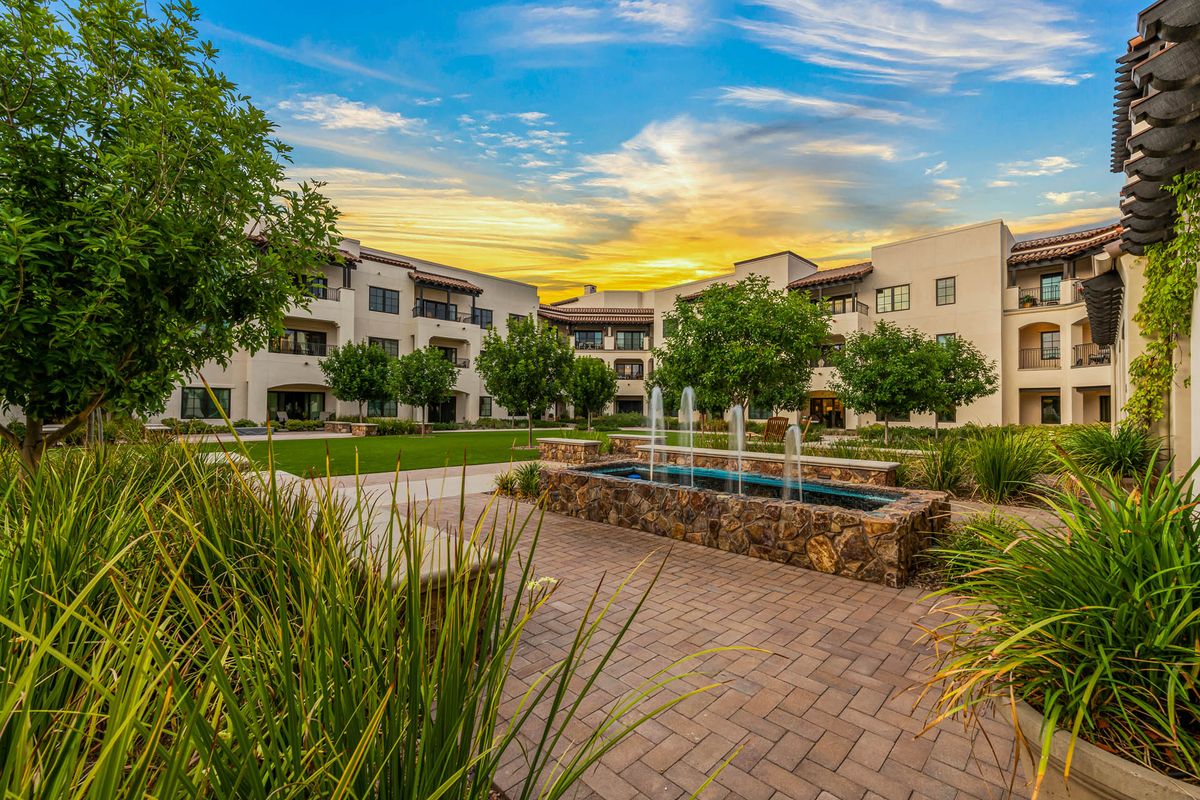 A serene view of apartment buildings with a central fountain and lush landscaping during sunset.
