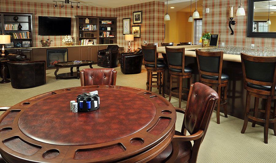A cozy game room with a poker table and leather chairs, featuring a bar area and bookshelf.