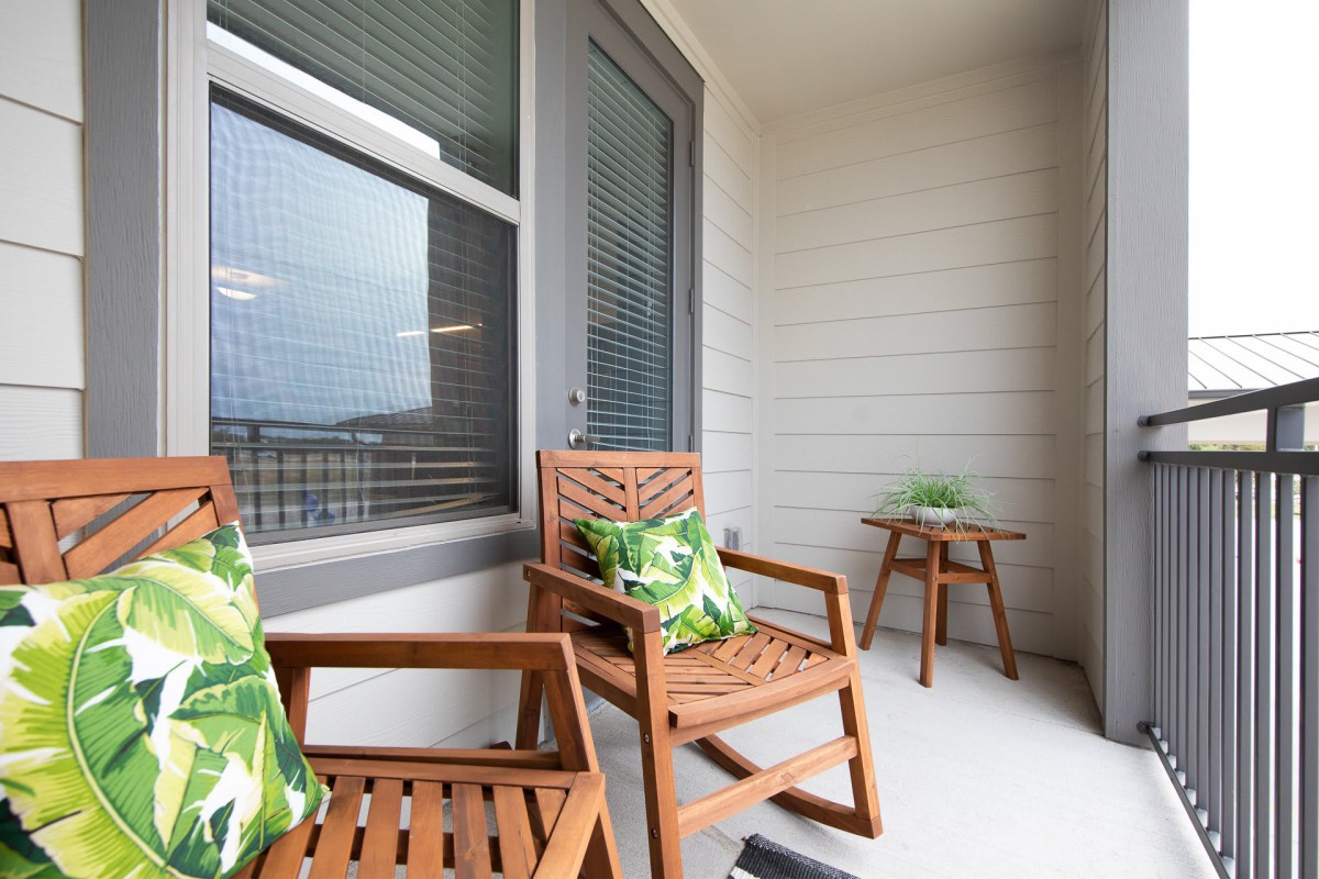 A cozy balcony setup with wooden chairs and a small table, featuring green patterned cushions and a potted plant.