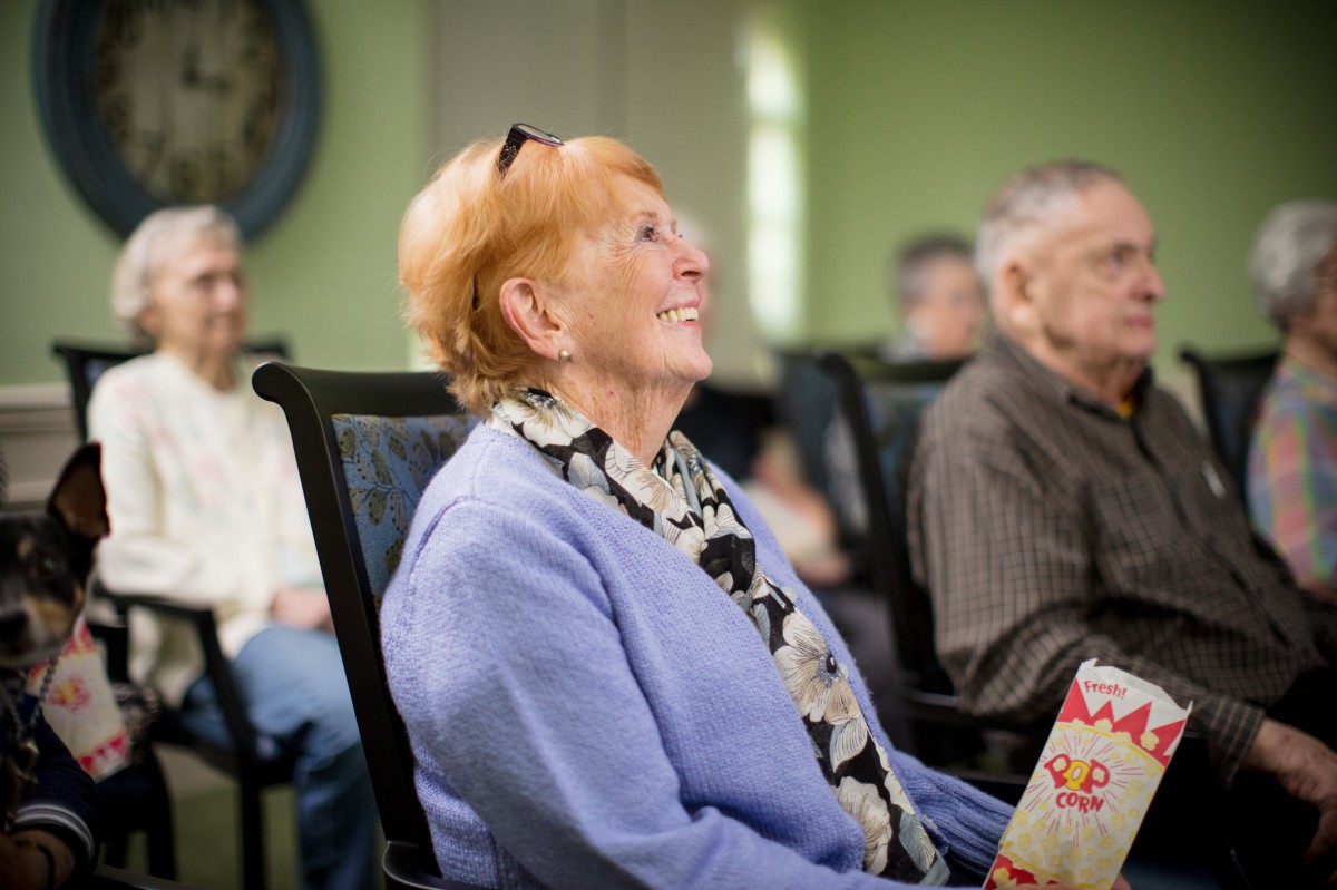 An elderly woman smiling while sitting in a group setting indoors.