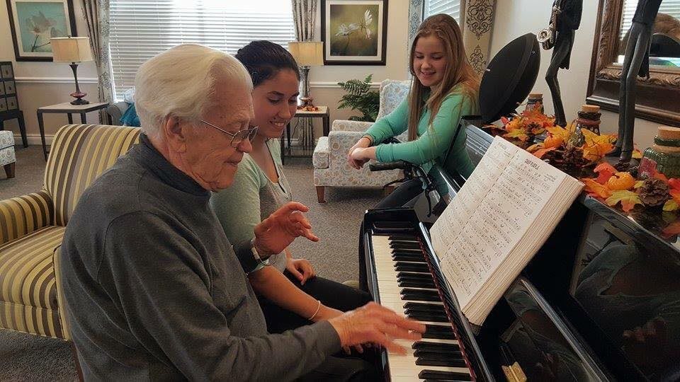 An elderly man teaching piano to two young women indoors.