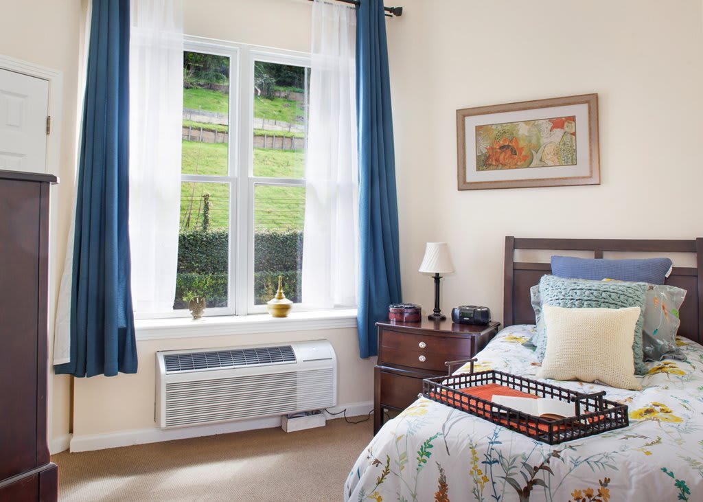 A cozy bedroom with a window view, featuring blue curtains and a neatly made bed.