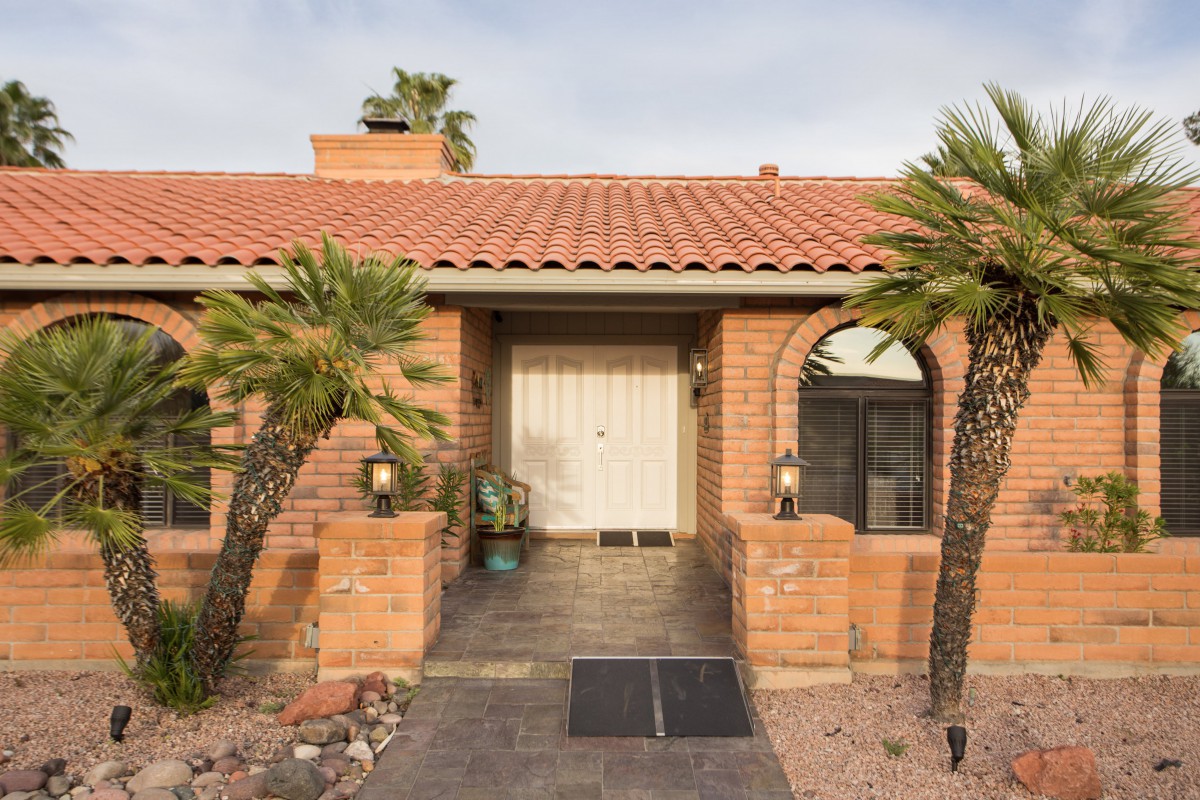 A single-story house with a brick exterior and desert landscaping, featuring palm trees and an arched window.