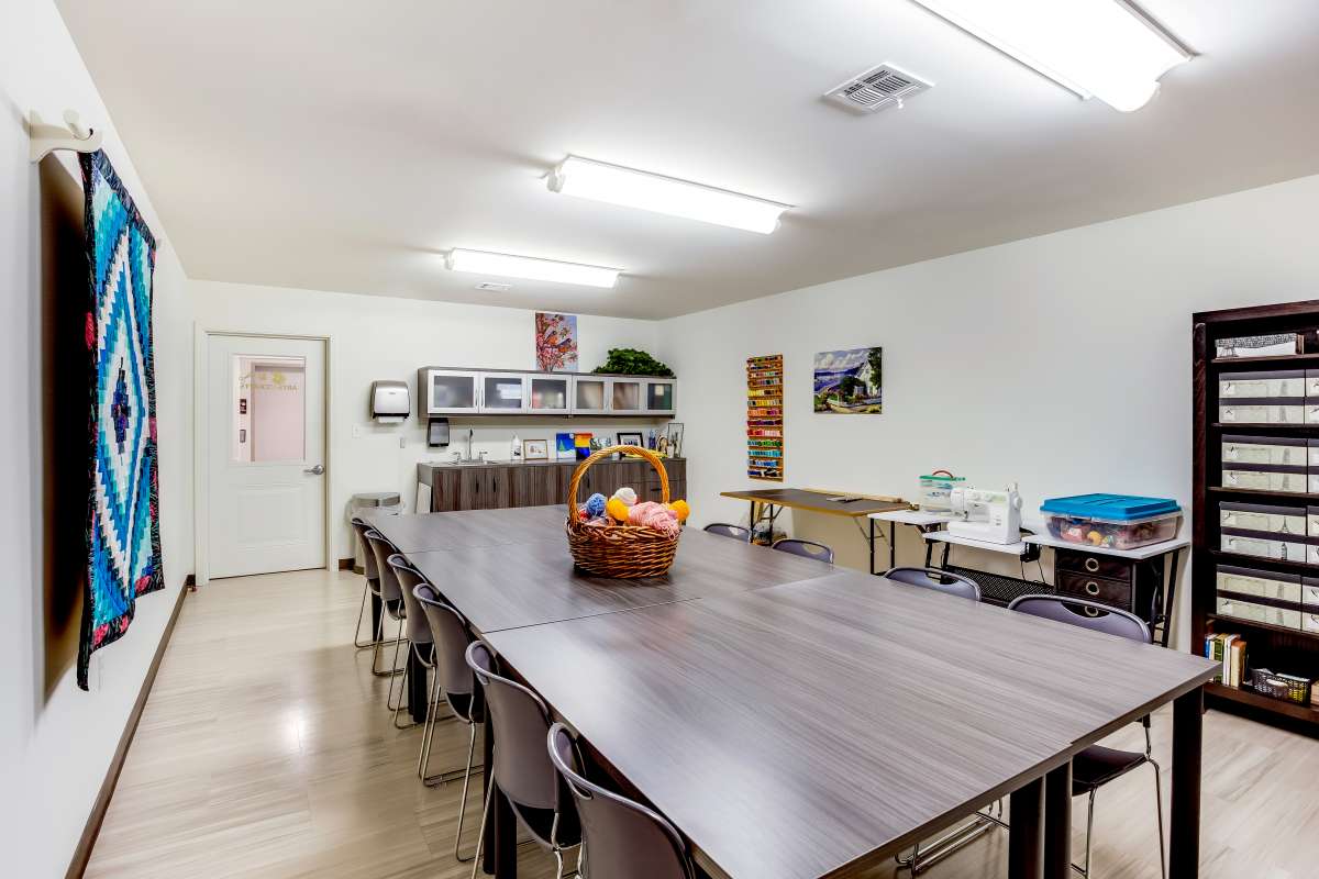 A spacious classroom or meeting room with a large table and chairs, featuring a whiteboard and shelves in the background.