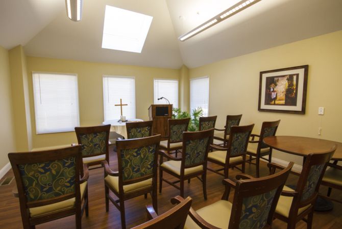 A small chapel with arranged chairs facing a pulpit and cross, illuminated by natural light from large windows and a skylight.