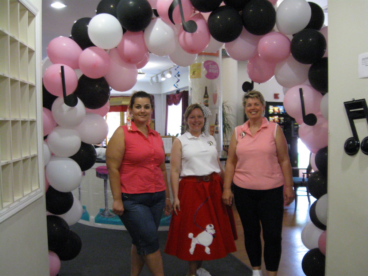 Three people standing under a colorful arch of pink, white, and black balloons, with one person wearing a poodle skirt.