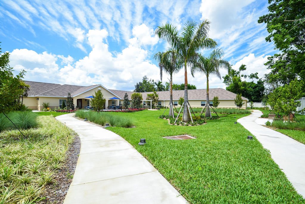 A scenic view of a house with a lush green lawn, palm trees, and a winding pathway under a partly cloudy sky.