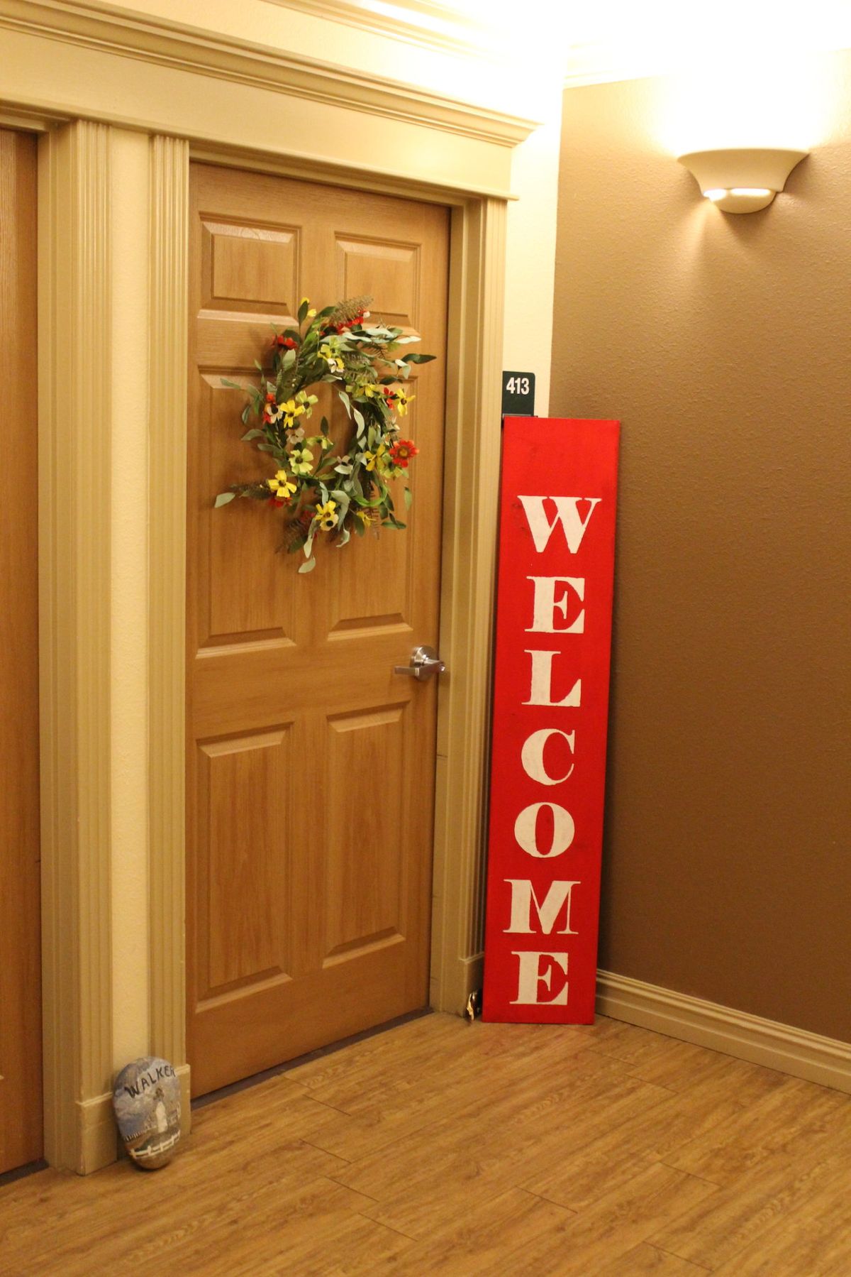 A wooden door with a floral wreath and a red welcome sign leaning against the wall.