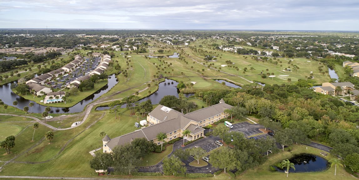 A scenic aerial view of a golf course with a clubhouse, surrounded by greenery and ponds.
