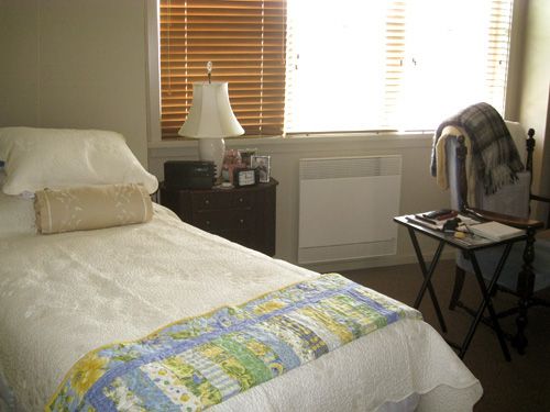 A cozy bedroom featuring a bed with a colorful quilt, a lamp on a nightstand, and a window with blinds.