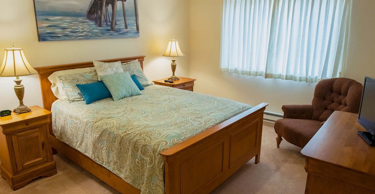 A cozy bedroom featuring a wooden bed with patterned bedding, a nightstand with a lamp, and a window with curtains.