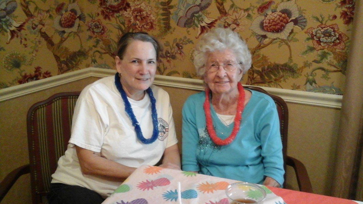 Two elderly women sitting together indoors, wearing colorful lei necklaces.