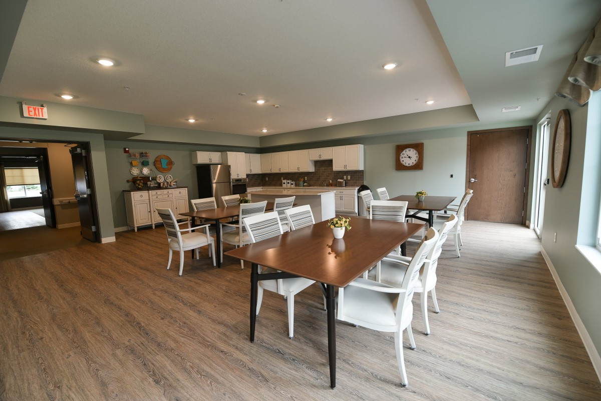 A modern dining room with wooden tables, white chairs, and a kitchen area in the background.