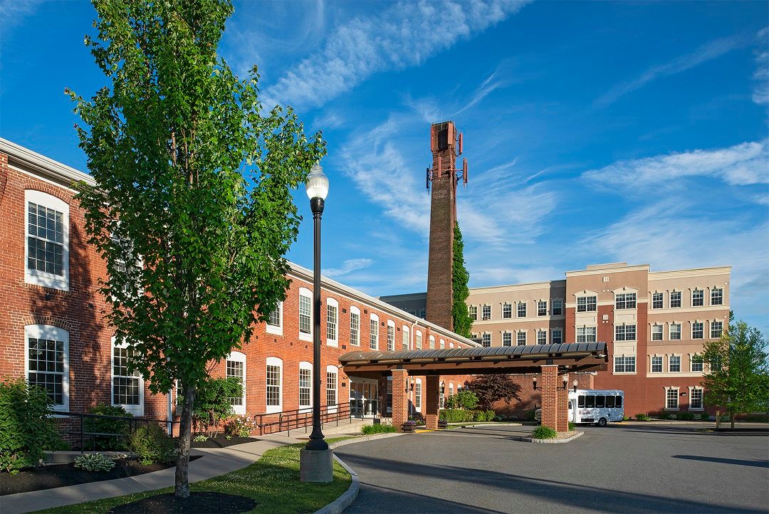 A brick building with a tall chimney stands against a blue sky, surrounded by trees and a lamp post.