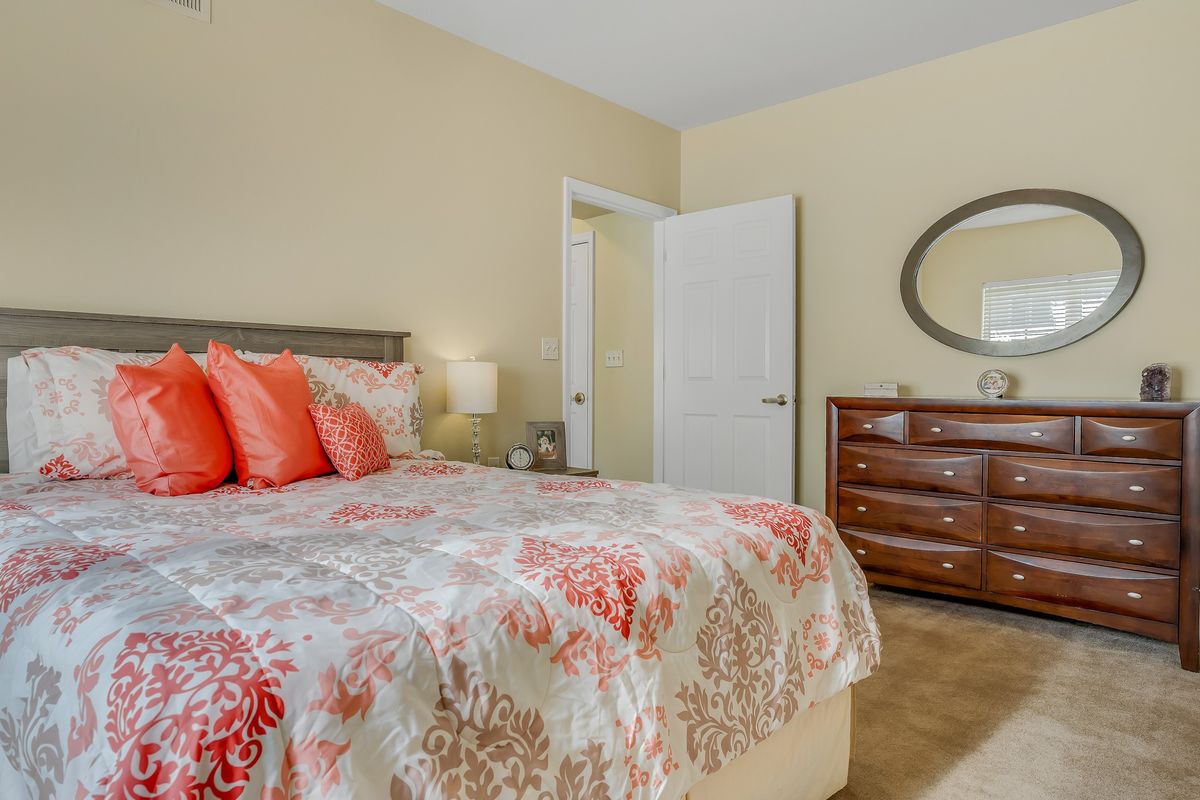 A cozy bedroom with a bed featuring floral bedding, a wooden dresser with an oval mirror, and soft lighting.