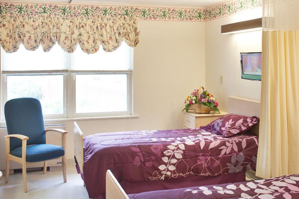 A cozy room with two beds featuring floral comforters, a window with curtains, and a small arrangement of flowers on a nightstand.
