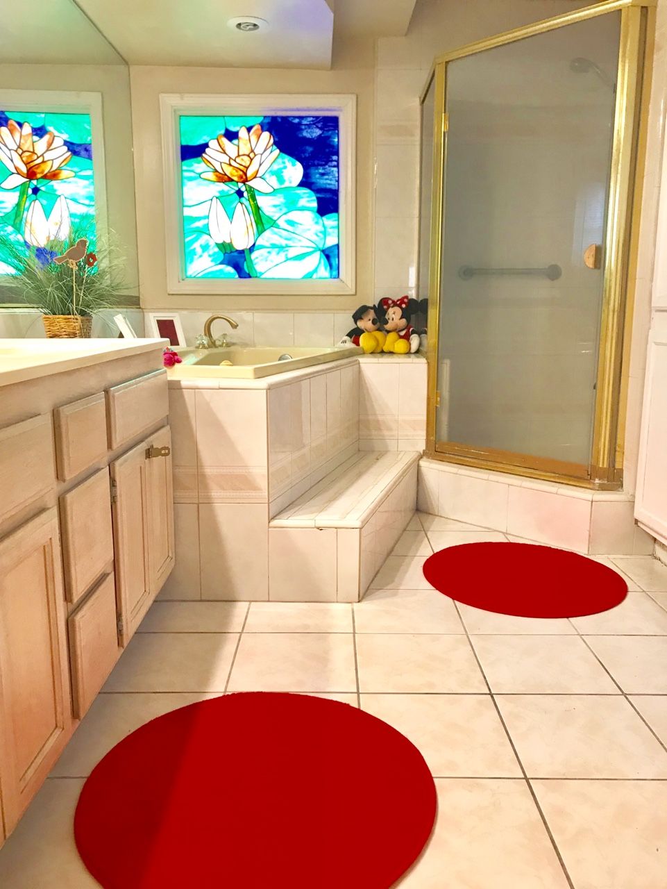 A well-lit bathroom featuring a bathtub, shower, and vibrant red rugs on a tiled floor, complemented by a stained glass window.
