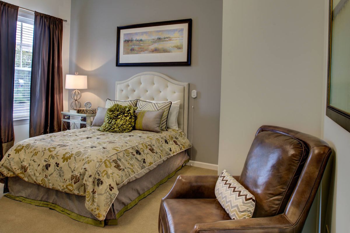A cozy bedroom featuring a plush bed with decorative pillows, an armchair, a nightstand with a lamp, and a framed painting on the wall.