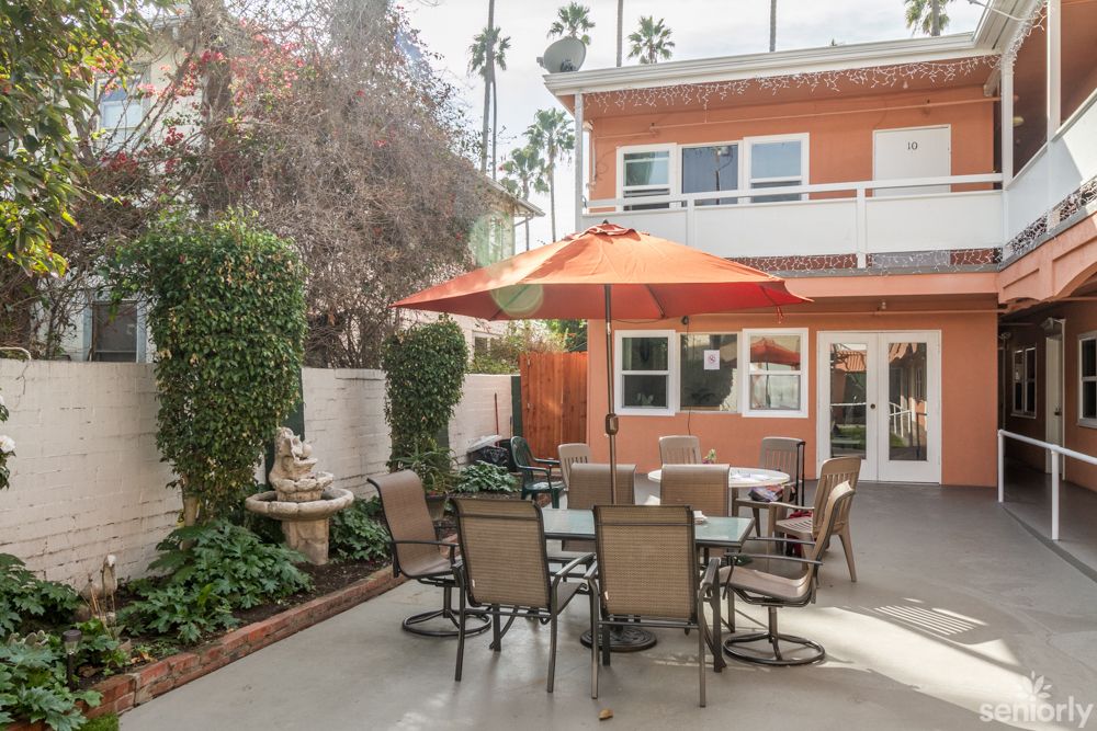 A cozy backyard patio with a table and chairs under an orange umbrella, surrounded by greenery.